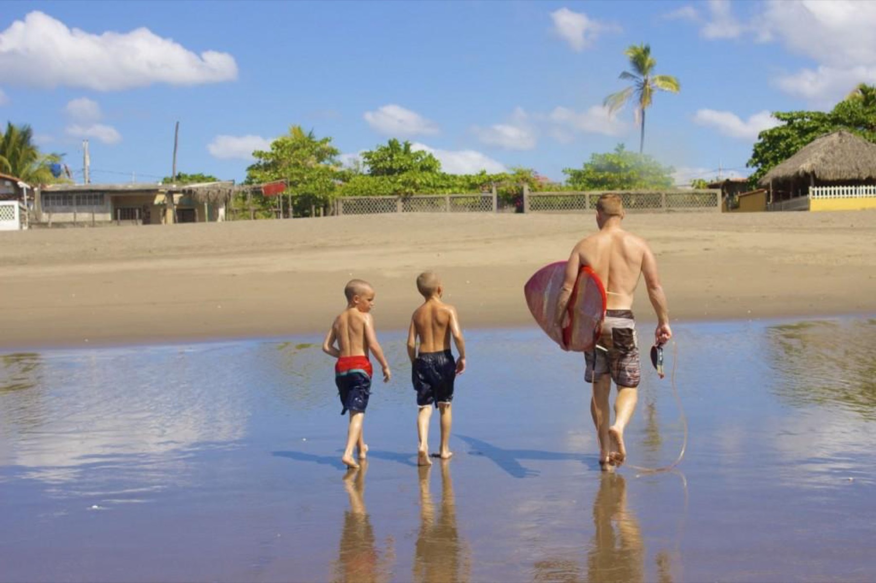 A father holds a surfboard beside his two children, with the beach town behind them