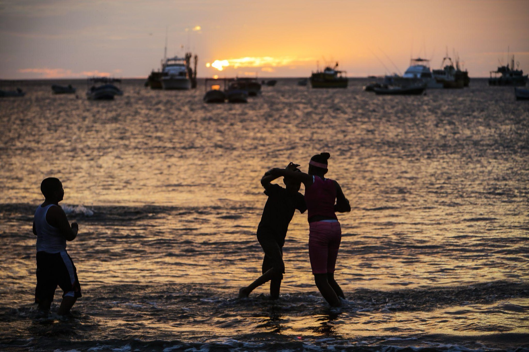 Three people wade into the bay at sunset, with the fishing boats of San Juan del Sur behind them